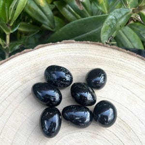 May include: A collection of seven polished black stones with smooth, rounded surfaces and subtle natural veining. The stones are arranged in a cluster, displaying a glossy, reflective finish that highlights their deep, dark colour and organic shapes.