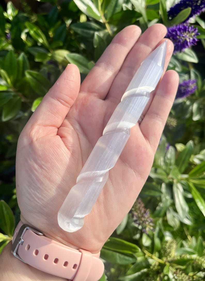 May include: A clear, spiraled selenite wand, held in a hand. The wand is translucent and has a smooth, polished appearance. The background features green foliage and purple flowers, creating a natural setting.