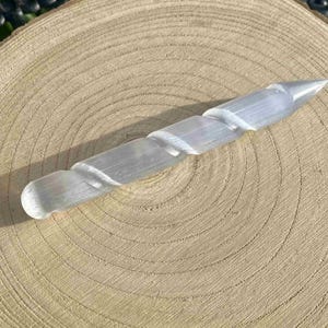 May include: A clear, spiral-carved selenite wand, approximately 6 inches long, rests on a light brown, circular wooden surface. The wand tapers to a point at one end and has a rounded tip on the other. The wood grain is visible.