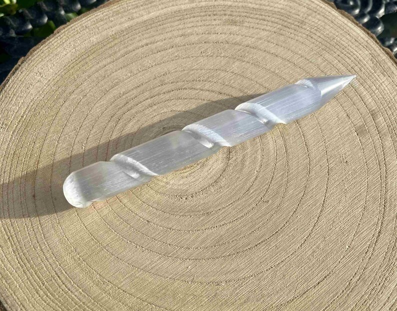 May include: A clear, spiral-carved selenite wand, approximately 6 inches long, rests on a light brown, circular wooden surface. The wand tapers to a point at one end and has a rounded tip on the other. The wood grain is visible.