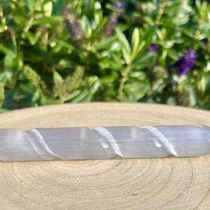 May include: A light gray selenite wand with a spiral carved design. The wand has a rounded end and tapers to a point. The wand is resting on a wooden surface, with green foliage in the background.