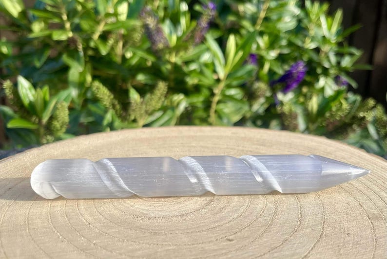 May include: A light gray selenite wand with a spiral carved design. The wand has a rounded end and tapers to a point. The wand is resting on a wooden surface, with green foliage in the background.