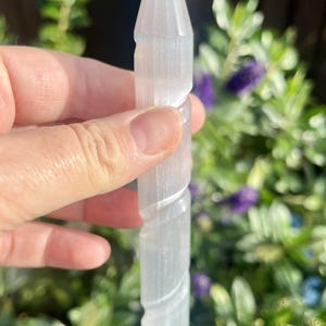 May include: A clear, elongated selenite crystal wand with a pointed tip and a spiraling carved design. The crystal is held against a backdrop of green foliage and purple flowers, creating a natural aesthetic.