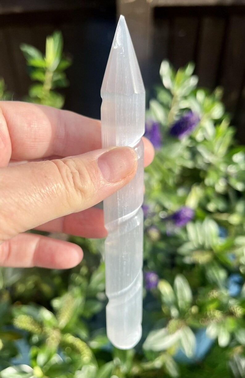 May include: A clear, elongated selenite crystal wand with a pointed tip and a spiraling carved design. The crystal is held against a backdrop of green foliage and purple flowers, creating a natural aesthetic.