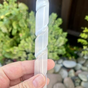 May include: A clear, white selenite crystal wand with a pointed tip and a spiral design. The wand is held in a hand against a blurred background of green foliage and stones. This crystal wand is a decorative item.