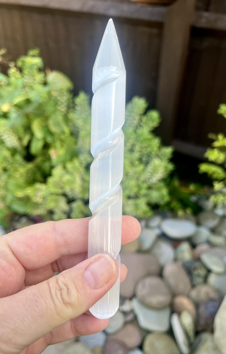 May include: A clear, white selenite crystal wand with a pointed tip and a spiral design. The wand is held in a hand against a blurred background of green foliage and stones. This crystal wand is a decorative item.