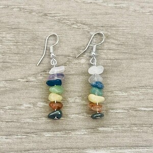 May include: A pair of silver-coloured drop earrings. Each earring has a vertical arrangement of polished gemstones in white, purple, blue, green, yellow, orange, and dark green. The earrings are set against a light wood grain background.
