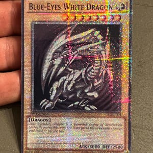 Puede incluir: Una tarjeta coleccionable holográfica con un dragón blanco. La tarjeta dice "BLUE-EYES WHITE DRAGON" en la parte superior. La tarjeta tiene el texto "ATK/3000 DEF/2500" en la parte inferior. Es un artículo de colección.