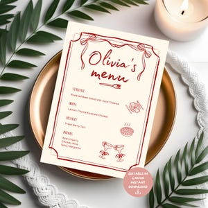 May include: A cream-colored menu with burgundy text and illustrations, titled "Olivia's menu." The menu lists starters, main courses, desserts, and drinks. The menu is on a gold plate, with greenery and a candle in the background.