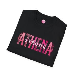 May include: Black t-shirt with the words "ATHENA STRONG" in a pink, decorative font. The text has a Celtic knot design. The shirt is folded and has a small pink tag on the upper right side.