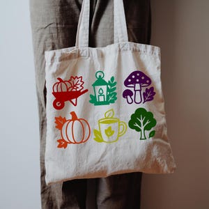 Könnte beinhalten: Eine cremefarbene Tragetasche mit bunten Herbstmotiven. Das Design umfasst einen Kürbis in einer Schubkarre, eine Laterne, einen Pilz, einen Kürbis, eine Teetasse und einen Baum. Die Tasche hat zwei Griffe.