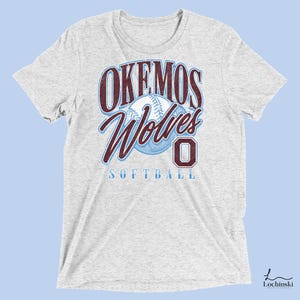 May include: A heather gray t-shirt featuring the text "Okemos Wolves Softball" in maroon and light blue. A baseball graphic is centered above the word "Softball". The number "0" is in maroon.