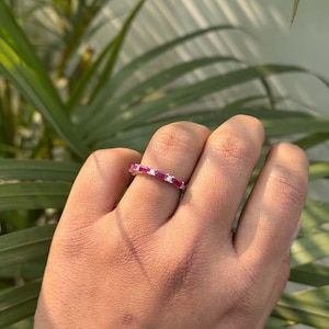 Op de afbeelding: Een zilverkleurige ring met een afwisselend patroon van rechthoekige baguette-geslepen roze robijnen en kleine ronde helderwitte stenen, gezet langs de bovenste helft van de ring voor een sprankelend en kleurrijk visueel effect.