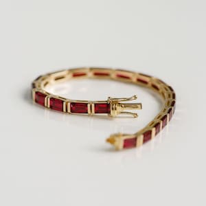 Könnte beinhalten: Ein goldenes Armband mit rechteckigen roten Edelsteinen. Das Armband ist geöffnet und zeigt den Verschlussmechanismus. Die Edelsteine sind in einem goldenen Band gefasst. Das Armband liegt auf einer weißen Oberfläche.