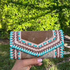 May include: A brown and tan leather wallet with turquoise stitching. The wallet has a flap closure and a decorative stitched design. The wallet is held in a hand against a background of green foliage.
