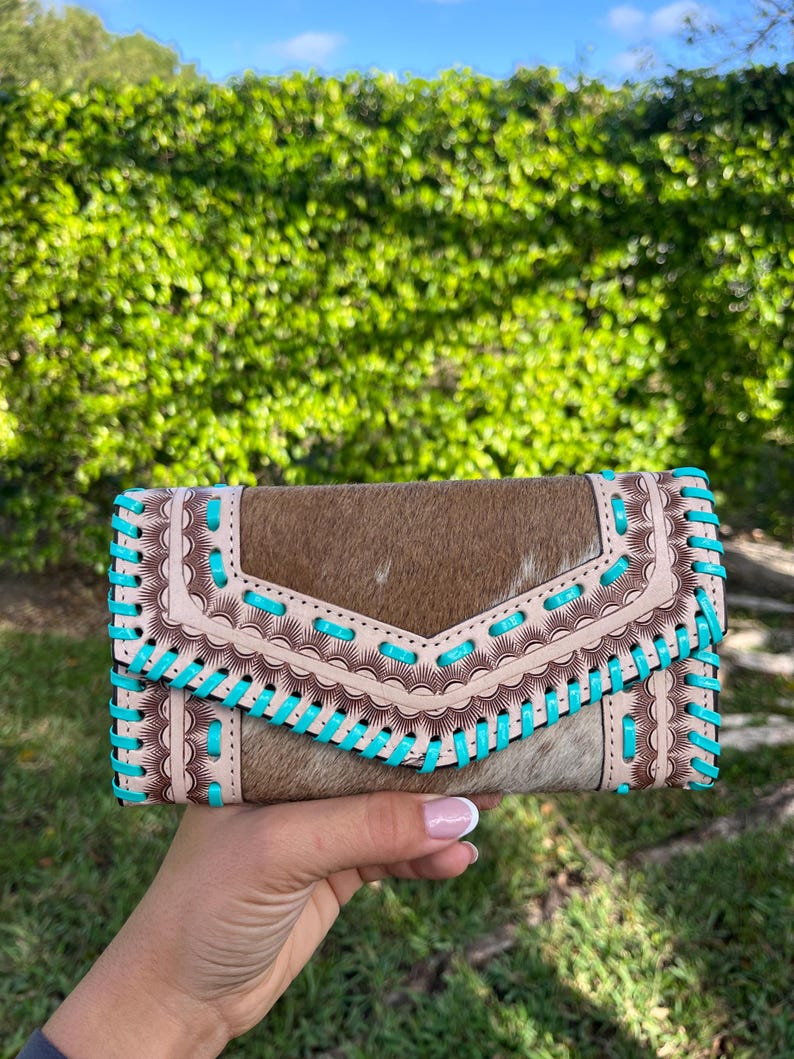 May include: A brown and tan leather wallet with turquoise stitching. The wallet has a flap closure and a decorative stitched design. The wallet is held in a hand against a background of green foliage.