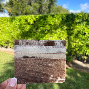 May include: A rectangular cardholder with a cowhide design. The cardholder features a mix of brown, white, and tan fur. The edges are stitched with brown thread. The cardholder is held in front of a green hedge.