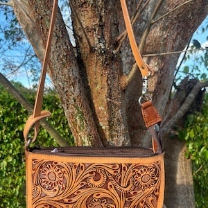 May include: A tan leather crossbody bag with a floral embossed design. The bag features a long, adjustable strap and a zipper closure. The intricate pattern includes stylized flowers and swirling leaves, with a darker brown border.
