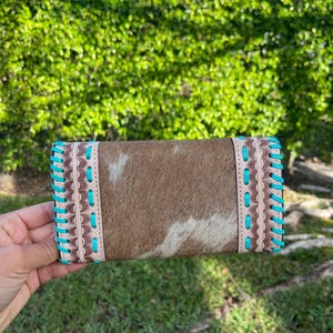 May include: A rectangular cowhide wallet with a brown and white pattern. The wallet has light brown leather trim and turquoise stitching along the edges. The wallet is held in front of a green hedge.