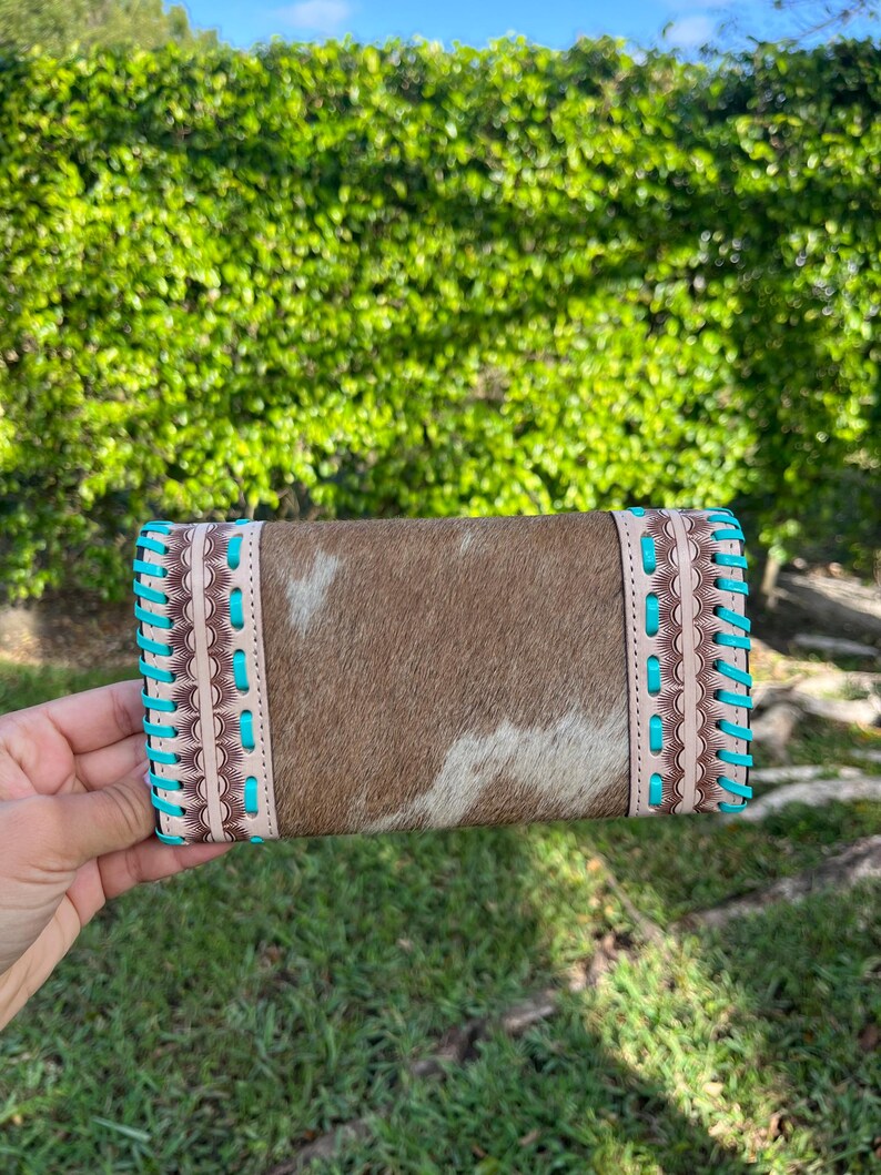 May include: A rectangular cowhide wallet with a brown and white pattern. The wallet has light brown leather trim and turquoise stitching along the edges. The wallet is held in front of a green hedge.