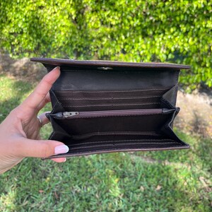 May include: Open brown leather wallet with multiple card slots and a zippered compartment. The wallet is held open, revealing the interior. The background is a blurred view of green foliage and grass.