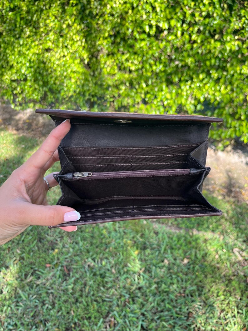 May include: Open brown leather wallet with multiple card slots and a zippered compartment. The wallet is held open, revealing the interior. The background is a blurred view of green foliage and grass.
