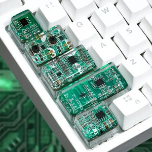 Può includere: Quattro tasti meccanici personalizzati per tastiera con circuiti stampati verdi a vista, componenti elettronici e microchip visibili. I tasti sono trasparenti, di forma rettangolare e posizionati accanto ai tasti bianchi standard.
