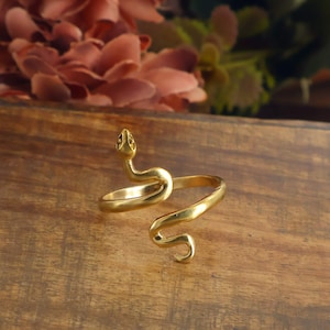 May include: A gold-toned metal ring shaped like a coiled snake. The ring features a detailed snake head at one end and a curved tail at the other, forming an open, adjustable band design with a smooth, polished finish throughout the entire piece.