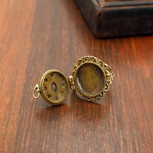 May include: Open antique brass locket ring with an oval setting and decorative filigree. The locket has a hinged compartment and a small loop for attaching a chain. The ring is on a wooden surface.