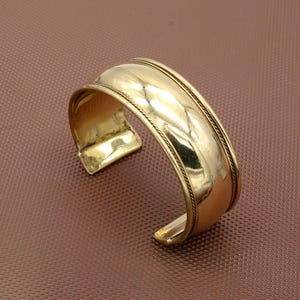 Op de afbeelding: Een goudkleurige manchetarmband met een breed, gebogen ontwerp. De armband heeft een centrale band met een getextureerd oppervlak, omlijst door twee dunnere, touwachtige banden. Het open ontwerp maakt het gemakkelijk om te dragen.
