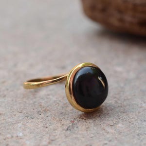 Puede incluir: Un anillo de oro con una piedra preciosa negra, grande y ovalada. La piedra preciosa está engastada en un bisel dorado, y la banda también es dorada. El anillo se muestra sobre una superficie neutra.
