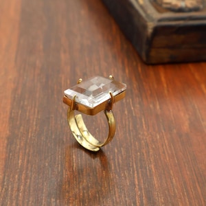 May include: A gold-toned ring with a large, rectangular, clear gemstone. The ring's band is adjustable, and the gemstone is set in a gold-colored bezel. The ring is displayed on a wooden surface.