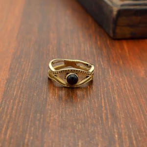 May include: A gold-colored ring with an eye-shaped design. The ring features a central, round, black stone and intricate detailing. The ring is set on a wooden surface.