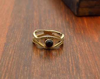 May include: A gold-colored ring with an eye-shaped design. The ring features a central, round, black stone and intricate detailing. The ring is set on a wooden surface.