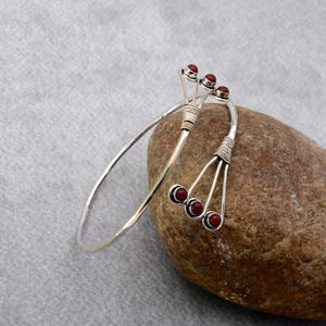 May include: A silver-toned cuff bracelet with three red circular accents on each of the two decorative ends. The bracelet is resting on a brown stone against a gray background. The ends of the bracelet have a wire-wrapped design.