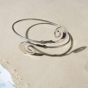 Pode incluir: Uma pulseira de braço prateada com um design em espiral em cada extremidade. A pulseira aberta tem um design simples e elegante. As espirais são lisas e arredondadas, e o metal tem um acabamento polido. A pulseira é provavelmente feita de um metal leve.