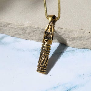 May include: A gold-colored pendant necklace with a cylindrical design. The pendant features intricate patterns, including a trident-like shape at the top and rope-like detailing. The necklace chain is a thin, gold-colored snake chain.