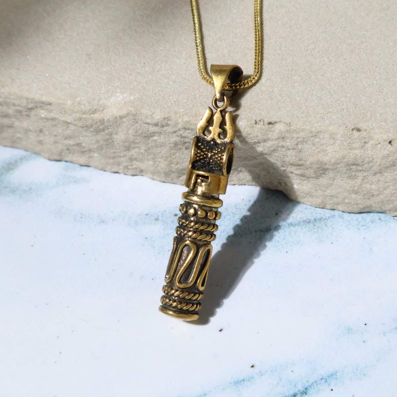 May include: A gold-colored pendant necklace with a cylindrical design. The pendant features intricate patterns, including a trident-like shape at the top and rope-like detailing. The necklace chain is a thin, gold-colored snake chain.