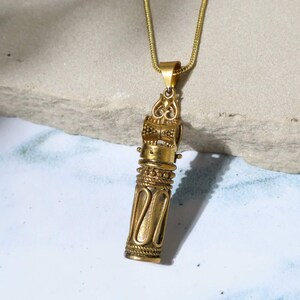 May include: A gold-colored pendant necklace with an ornate, cylindrical design. The pendant features intricate detailing, including a decorative top and a textured body. The necklace chain is also gold-toned, creating a cohesive look. The pendant hangs against a neutral background.