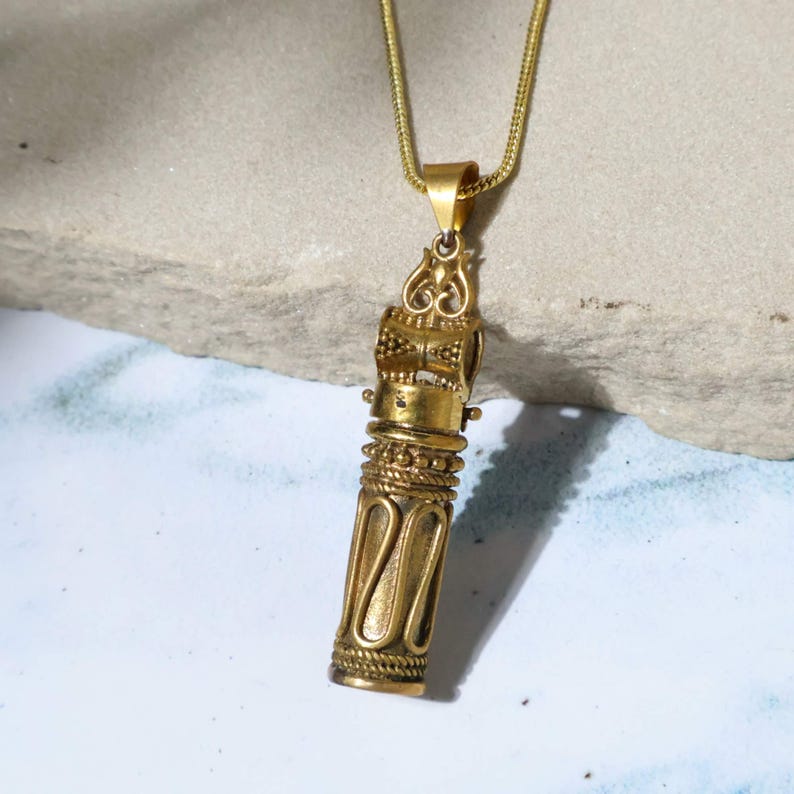 May include: A gold-colored pendant necklace with an ornate, cylindrical design. The pendant features intricate detailing, including a decorative top and a textured body. The necklace chain is also gold-toned, creating a cohesive look. The pendant hangs against a neutral background.