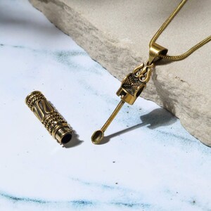 May include: A gold-colored pendant necklace with a detailed design, featuring a small spoon-like tool and a cylindrical container. The necklace is displayed on a light-colored stone surface, showcasing the intricate metalwork and craftsmanship.