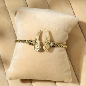 May include: Gold-coloured bracelet with a wing design. The bracelet has a twisted rope-like band with detailed wing accents. The wings are positioned at the centre of the bracelet, creating a unique and stylish accessory.