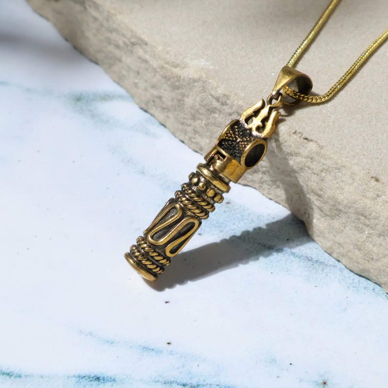 May include: A gold-colored pendant necklace with an ornate, cylindrical pendant. The pendant features intricate designs and a trident-like symbol at the top. The necklace chain is also gold-colored, and the pendant is displayed against a light-colored stone.