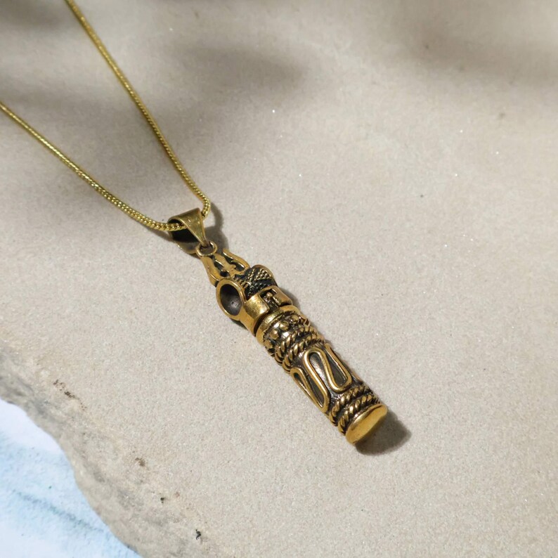 May include: A gold-colored pendant necklace with an ornate, cylindrical design. The pendant features intricate patterns and is suspended from a delicate gold chain. The necklace is displayed on a neutral-toned surface.