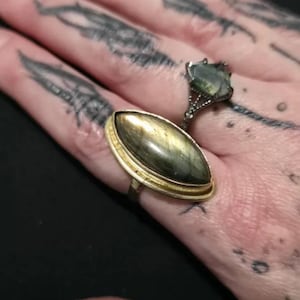 May include: A gold-toned ring featuring a large, elongated marquise-cut labradorite stone with iridescent grey and gold flashes, paired with a smaller, dark metal ring set with a faceted, shield-shaped grey stone and intricate metal detailing.