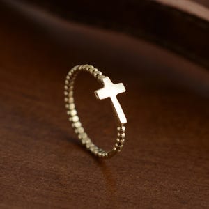 May include: A gold-colored ring with a cross design. The ring band is made of small, round beads. The cross is a solid, simple shape. The ring is set against a brown background.