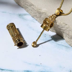 May include: A gold-colored pendant necklace with a small, ornate container and a spoon-like tool. The container is cylindrical with decorative detailing. The spoon is attached to a crown-shaped element, and the necklace chain is visible.
