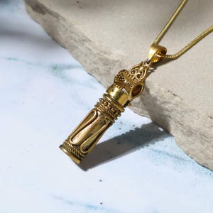 May include: A gold-colored pendant necklace with an ornate, cylindrical design. The pendant features intricate detailing and is suspended from a delicate gold chain. The necklace is displayed against a neutral background, highlighting its vintage aesthetic.