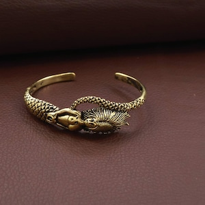 Pode incluir: Uma pulseira de metal em tom dourado com um design de sereia esculpido. A pulseira apresenta um padrão de escamas texturizado ao longo da banda, com o corpo e o cabelo fluido da sereia formando o ponto focal central da peça aberta.