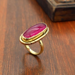 May include: A gold-coloured ring featuring a large, oval magenta gemstone. The ring has a thick band and a decorative setting. The ring is displayed on a wooden surface.